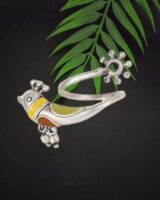 Colorful traditional Indian jewelry piece with yellow, green, and orange accents, featuring a cat motif and intricate details, placed on a dark background with green leaves for contrast.