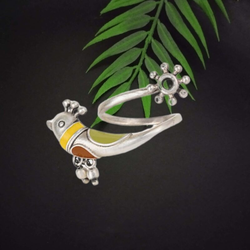 Colorful traditional Indian jewelry piece with yellow, green, and orange accents, featuring a cat motif and intricate details, placed on a dark background with green leaves for contrast.