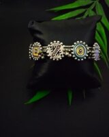 Silver traditional Indian jewelry bracelet with colorful enamel accents, intricate design, and beaded details, displayed on a black satin cushion with green leaves in the background.