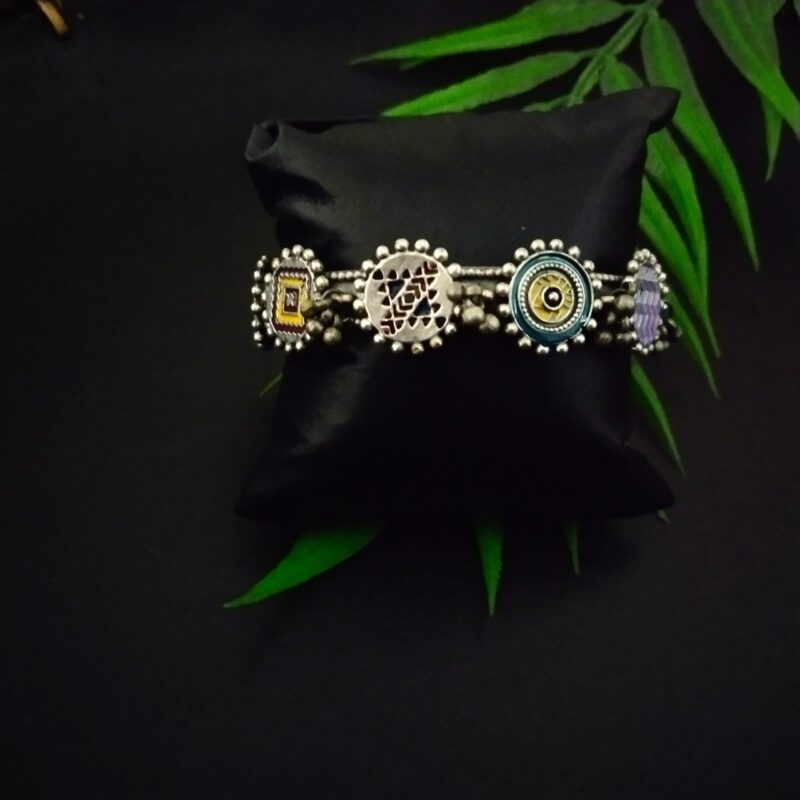 Silver traditional Indian jewelry bracelet with colorful enamel accents, intricate design, and beaded details, displayed on a black satin cushion with green leaves in the background.