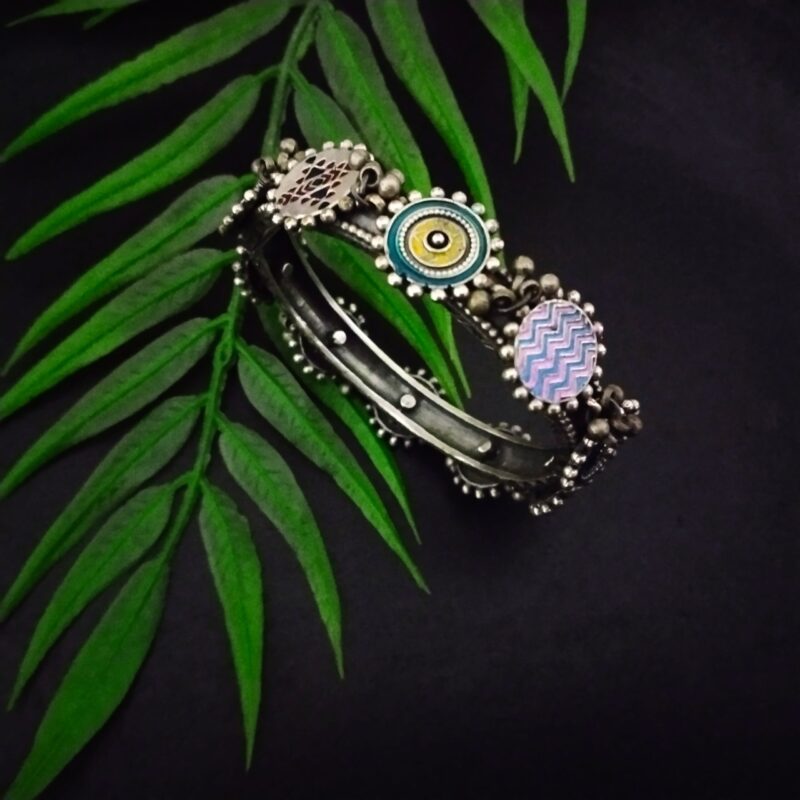 Intricate Indian boho silver bracelet featuring colorful enamel and geometric designs, showcased on a green fern leaf against a black background, highlighting traditional ethnic jewelry craftsmanship.