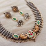 Intricate handcrafted Indian fusion jewelry set including necklace and earrings with peacock motifs, green and red stones, pearls, and oxidized silver finish, perfect for traditional festivals and celebrations.