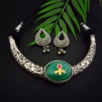 Intricate silver necklace with green pendant featuring a pink stone and golden design, paired with matching silver jhumka earrings with floral motifs and dangling bells, displayed on a dark background with green leaves.