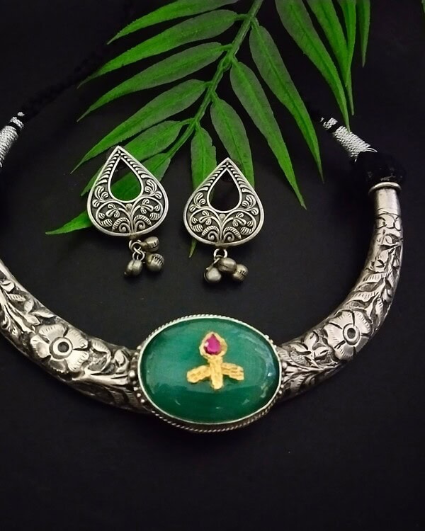 Intricate silver necklace with green pendant featuring a pink stone and golden design, paired with matching silver jhumka earrings with floral motifs and dangling bells, displayed on a dark background with green leaves.