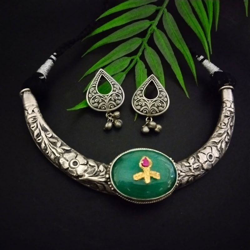Intricate silver necklace with green pendant featuring a pink stone and golden design, paired with matching silver jhumka earrings with floral motifs and dangling bells, displayed on a dark background with green leaves.