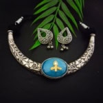 Elegant Indian jewelry set featuring a silver necklace with floral engravings and a central blue gemstone pendant, paired with matching silver earrings with intricate designs and dangling beads, displayed with green leaves on a black background.