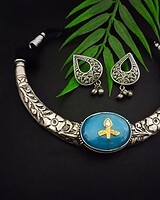 Elegant Indian jewelry set featuring a silver necklace with floral engravings and a central blue gemstone pendant, paired with matching silver earrings with intricate designs and dangling beads, displayed with green leaves on a black background.