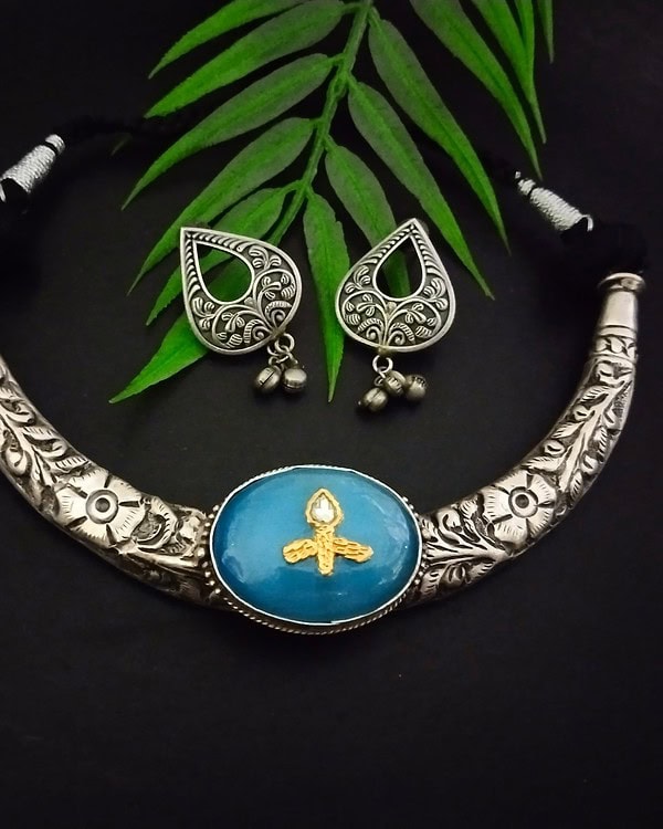 Elegant Indian jewelry set featuring a silver necklace with floral engravings and a central blue gemstone pendant, paired with matching silver earrings with intricate designs and dangling beads, displayed with green leaves on a black background.