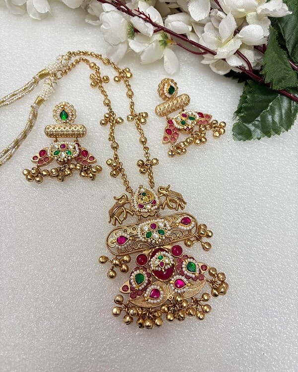 Intricate gold jewelry set featuring colorful stones including green, pink, and white, with matching earrings and necklace, displayed on a white surface with floral accents. Perfect for traditional Indian occasions.
