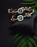 Silver and colorful traditional Indian jewelry set including bangles and earrings with turquoise, red, and green stones, displayed on a black silk cushion with green leaves backdrop.