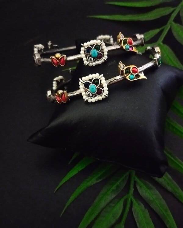 Silver and colorful traditional Indian jewelry set including bangles and earrings with turquoise, red, and green stones, displayed on a black silk cushion with green leaves backdrop.
