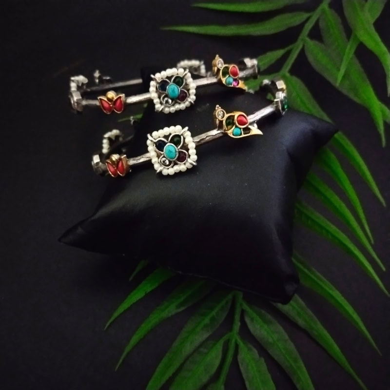 Silver and colorful traditional Indian jewelry set including bangles and earrings with turquoise, red, and green stones, displayed on a black silk cushion with green leaves backdrop.