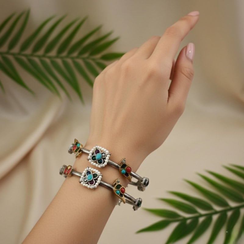 Hand wearing traditional Indian silver bangles with colorful inlay accents and intricate design, styled with a neutral background and green leaves to showcase elegant jewelry.