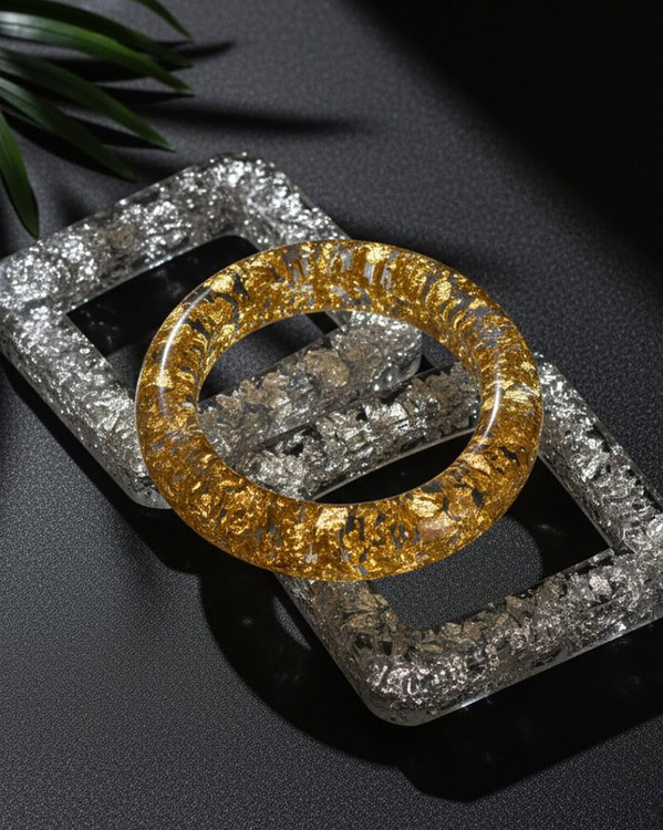 Shimmering gold and silver resin bangles and rectangular futuristic jewelry display tray on black textured background with green leaf accent.