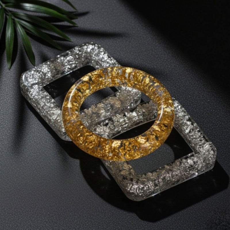Shimmering gold and silver resin bangles and rectangular futuristic jewelry display tray on black textured background with green leaf accent.