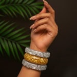 Vibrant Indian fashion jewelry bangles on a stylish hand against a dark background with green leaves, showcasing a mix of silver and gold-toned accessories perfect for traditional or contemporary wear.
