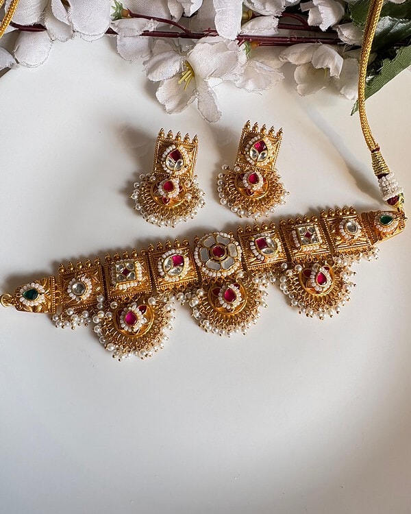 Intricate gold plated jewelry set including a choker necklace and matching earrings with red, white, and green stones, embellished with pearl accents, perfect for traditional Indian festivities and weddings.