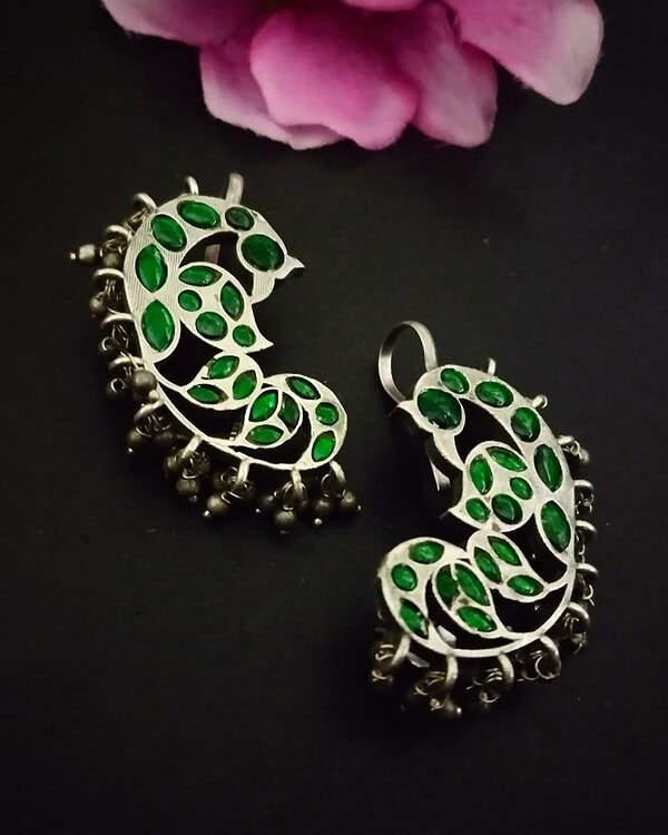 Shimmering silver earrings with green gem accents, featuring intricate leaf motifs and dangling embellishments, perfect for traditional Indian jewelry styles.