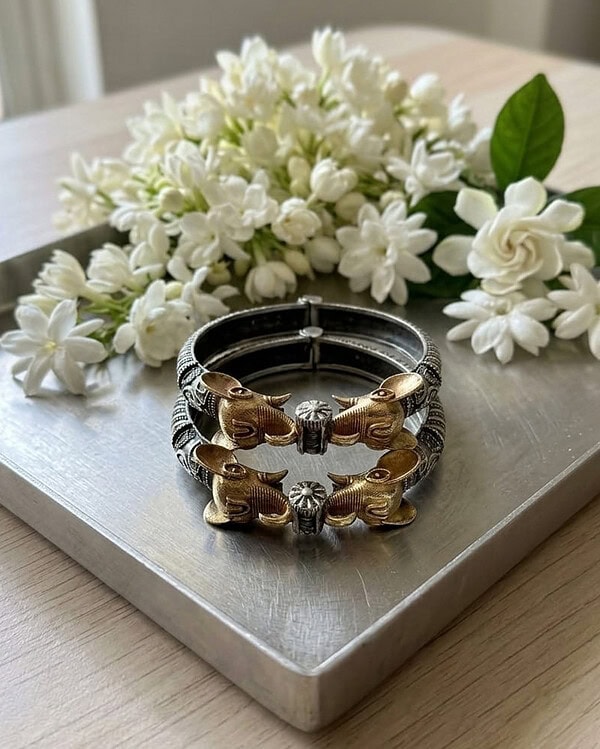 Beautiful gold and silver bracelet featuring intricate animal motifs, displayed on a neutral surface with a backdrop of fresh white flowers, perfect for Indian fashion jewelry lovers.